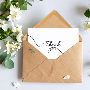 May include: A brown paper envelope contains a white card with the words "Thank you" in elegant script. A black line art design accompanies the text. White flowers and greenery are arranged around the envelope on a white surface.