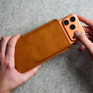 Peut inclure: Une pochette de téléphone en cuir beige avec des coutures visibles sur les bords, contenant partiellement un smartphone orange métallique doté d'un triple objectif photo à l'arrière. La pochette est rectangulaire avec des coins arrondis.