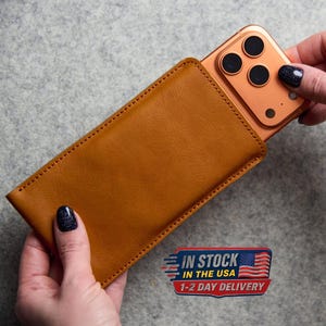 May include: A tan leather phone sleeve with visible stitching along the edges, holding a rose gold smartphone with a triple-lens camera system. The sleeve is rectangular and smooth, providing a snug fit for the device shown in the image.