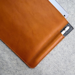May include: A brown leather tablet sleeve with a stitched border. A white stylus is in a side pocket. A silver tablet is partially visible inside the sleeve. The sleeve is on a gray surface.