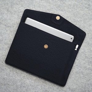 May include: A dark navy blue leather tablet sleeve with a gold snap closure. The sleeve is open, revealing a silver tablet inside. A white stylus is visible in a small pocket. The leather has a textured appearance.