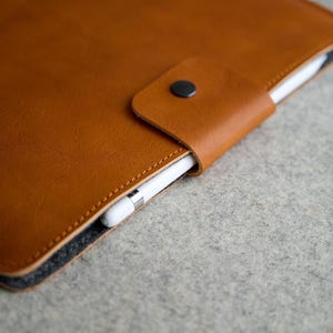 Op de afbeelding: Close-up van een bruine leren tablethoes met een zwarte knoopsluiting. Een witte stylus wordt in een lus op de hoes gehouden. De hoes ligt op een grijze ondergrond.