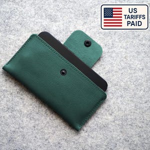 May include: A dark green leather phone case with a snap closure, holding a black smartphone. The case is rectangular and has a flap that folds over to secure the phone. The text "US Tariffs Paid" is in the upper right corner.
