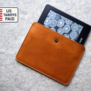 May include: A Kindle e-reader in a brown leather case. The e-reader is black with a screen displaying a pattern. The case has a snap closure. A sticker with the US flag and text "US Tariffs Paid" is in the upper left corner.