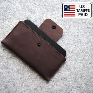 May include: A brown leather phone case with a snap closure, holding a black smartphone. The case is open, revealing the phone. The image includes a small sign with the US flag and the text "US Tariffs Paid."