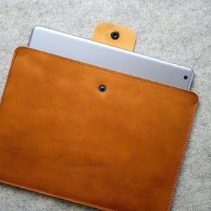 May include: A tan leather tablet case with a silver tablet partially inserted. The case has a snap closure and a stitched border. The tablet is visible at the top, showing its screen and edges. The case is made of a smooth, textured leather.