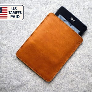 Può includere: Una custodia in pelle marrone chiaro per Kindle con un dispositivo Kindle all'interno. La custodia è rettangolare con angoli arrotondati e bordi cuciti. Il Kindle è nero con il marchio "Kindle" visibile. Lo sfondo è una superficie grigio chiaro.