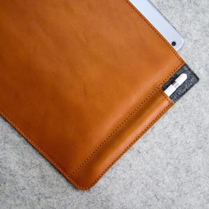 Puede incluir: Una funda de tableta de cuero marrón claro con un bolsillo de fieltro gris para un lápiz óptico. La funda está cosida a lo largo de los bordes y contiene una tableta plateada. El cuero tiene una textura suave y un color cálido y natural.
