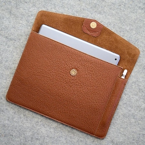 Könnte beinhalten: Eine braune Tablet-Hülle aus Leder mit strukturierter Oberfläche. Die Hülle ist geöffnet und zeigt ein silbernes Tablet und einen weißen Stift. Sie hat einen Druckknopfverschluss und ein weiches, braunes Innenfutter.