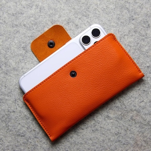 May include: Orange leather phone case with a snap closure, holding a white smartphone. The case has a textured surface and a contrasting tan leather flap. The phone's camera lenses are visible.