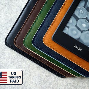Pode incluir: Uma pilha de capas de couro para leitores de e-books Kindle em várias cores: preto, castanho, verde, azul marinho e bege. O dispositivo Kindle é visível, exibindo uma tela. As capas têm bordas costuradas. A imagem inclui um adesivo "US Tariffs Paid".