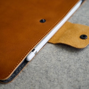 Puede incluir: Primer plano de una funda de tableta de cuero marrón con un lápiz blanco insertado. La funda tiene una solapa con cierre de botón a presión. Las costuras son visibles a lo largo de los bordes. El fondo es una superficie gris.
