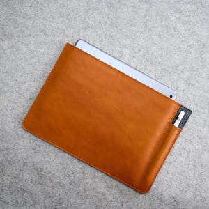 Könnte beinhalten: Eine rechteckige, hellbraune Tablet-Hülle aus Leder mit einer grauen Filztasche und einem teilweise eingesetzten silbernen Tablet. Ein weißer Stift befindet sich in der Tasche. Die Hülle liegt auf einer grauen Oberfläche.