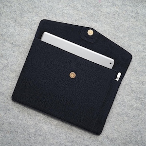 May include: A black leather tablet sleeve with a gold popper closure. The sleeve is open, revealing a silver tablet and a white stylus. The leather has a textured appearance, and the background is a light grey.