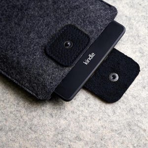 Pode incluir: Um leitor de e-books Kindle preto parcialmente inserido em uma capa de feltro cinza escuro. A capa tem uma aba de couro preta com fecho de pressão. A palavra "Kindle" é impressa em branco no dispositivo.