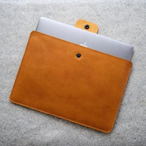 May include: A tan leather laptop sleeve featuring a rectangular shape with rounded corners and a single snap button closure. A silver laptop with a visible apple logo is partially inserted into the sleeve, which has a matching leather tab.