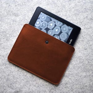 Può includere: Una custodia in pelle marrone per Kindle con un e-reader nero parzialmente visibile. La custodia ha una chiusura a scatto e un bordo cucito. L'e-reader mostra un'immagine in scala di grigi. La custodia è su una superficie grigia.