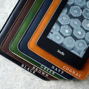 Pode incluir: Uma pilha de cinco capas de couro para e-reader nas cores preta, marrom, verde, azul marinho e conhaque, com um e-reader Kindle preto em cima. Cada capa apresenta costuras visíveis nas bordas e o nome da cor em texto branco na base.