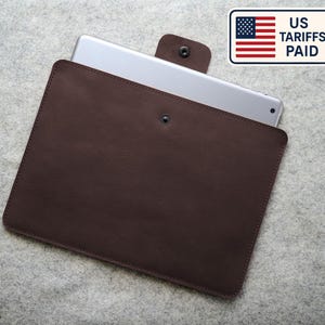 May include: A dark brown leather tablet sleeve with a snap closure, holding a silver tablet. The sleeve is rectangular and has a smooth texture. The image includes a US tariffs paid sticker.