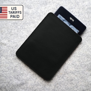 Op de afbeelding: Zwarte Kindle e-reader in een zwarte leren hoes. De Kindle is gedeeltelijk zichtbaar, met het Kindle-logo in het wit. De hoes is rechthoekig met afgeronde hoeken. Een sticker met de Amerikaanse vlag en de tekst "US Tariffs Paid" is zichtbaar.