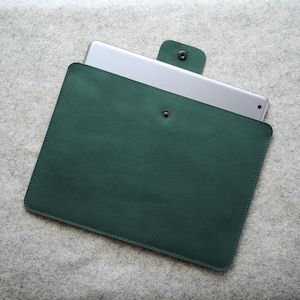 May include: A dark green leather tablet sleeve with a snap closure. The sleeve is rectangular and holds a silver tablet. The tablet is partially visible, and the sleeve is closed with a button clasp. The background is a light gray felt.