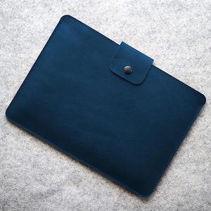 May include: A navy blue leather tablet case with a flap and a metal popper. The rectangular case has rounded corners and is stitched around the edges. It rests on a light grey felt surface.