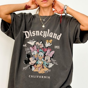Puede incluir: Camiseta gris oscuro con el logotipo de Disneyland, el texto "est. 1955" y un gr&aacute;fico de personajes de Disney. La palabra "CALIFORNIA" est&aacute; impresa debajo del gr&aacute;fico. La camiseta tiene mangas cortas.