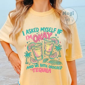 以下が含まれることがあります： バターイエローのTシャツ。水色とピンクで「I asked myself if I'm okay」と書かれたグラフィック、テキーラショットグラス2つとヤシの木が描かれています。「and we both ordered tequila」の文字が下にあります。