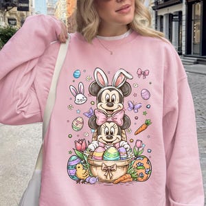 Puede incluir: Sudadera rosa claro con un dise&ntilde;o de Mickey y Minnie Mouse como conejos de Pascua, rodeados de huevos de Pascua, zanahorias y flores. La palabra "PINK" est&aacute; impresa en la parte inferior izquierda de la sudadera.