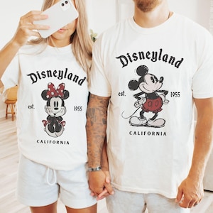 Puede incluir: Dos camisetas blancas con el logotipo de Disneyland y gr&aacute;ficos de Mickey y Minnie Mouse. Las camisetas tienen el texto "est. 1955 CALIFORNIA". La persona de la izquierda lleva pantalones cortos grises, y la persona de la derecha lleva pantalones cortos grises y calcetines naranjas.