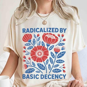 Op de afbeelding: Ivoor kleurig T-shirt met de tekst "RADICALIZED BY BASIC DECENCY" in blauw. Het shirt heeft een bloemenontwerp in rood en blauw. Het shirt heeft een relaxte pasvorm.