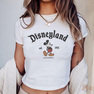 May include: White crop top featuring the Disneyland logo with Mickey Mouse. The design includes the text "Disneyland est. 1955 CALIFORNIA" in black. The shirt is a classic, casual style.