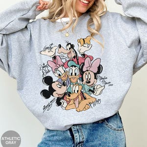 K&ouml;nnte beinhalten: Graues Sweatshirt mit einem farbenfrohen Aufdruck von Disney-Figuren, darunter Micky Maus, Minnie Maus, Donald Duck, Daisy Duck, Goofy und Pluto. Das Sweatshirt hat die Aufschrift "Mickey Mouse" und "Minnie Mouse".