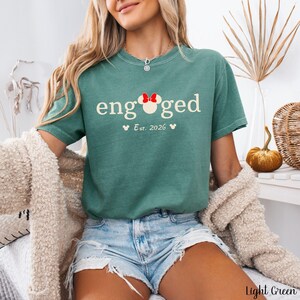 K&ouml;nnte beinhalten: Hellgr&uuml;nes T-Shirt mit dem Wort "engaged" in Wei&szlig;, mit einer Minnie-Mouse-Schleife. Darunter steht "Est. 2026" mit Mickey-Mouse-K&ouml;pfen. Das Shirt wird mit Jeansshorts kombiniert.