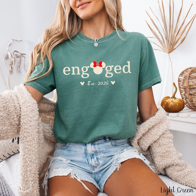 K&ouml;nnte beinhalten: Hellgr&uuml;nes T-Shirt mit dem Wort "engaged" in Wei&szlig;, mit einer Minnie-Mouse-Schleife. Darunter steht "Est. 2026" mit Mickey-Mouse-K&ouml;pfen. Das Shirt wird mit Jeansshorts kombiniert.