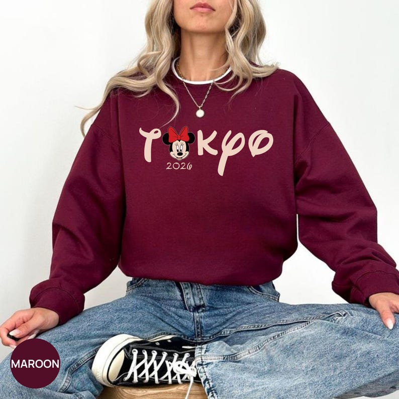 May include: Maroon sweatshirt with the word "Tokyo" in a stylized font, featuring a Minnie Mouse bow and the year "2026". The sweatshirt is a solid maroon color. The model is wearing blue jeans and black and white sneakers.