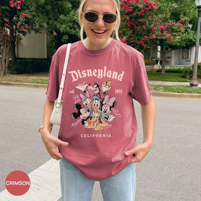 Puede incluir: Camiseta de color carmes&iacute; con la palabra "Disneyland" y un gr&aacute;fico de personajes de Disney. La camiseta incluye el texto "est. 1955" y "CALIFORNIA". Es de manga corta y cuello redondo.