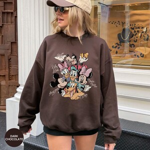K&ouml;nnte beinhalten: Dunkel schokoladenbraunes Sweatshirt mit Rundhalsausschnitt und langen &Auml;rmeln. Es zeigt eine farbenfrohe Grafik von Disney-Figuren wie Mickey und Minnie Mouse, Donald Duck und Pluto. Der Text "Dark Chocolate" befindet sich unten links.