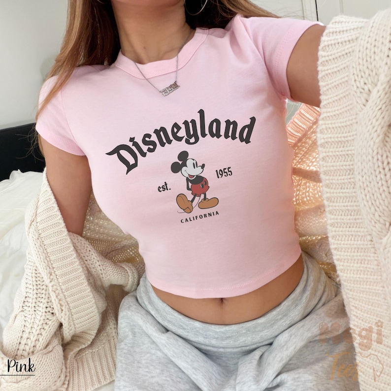 May include: Pink crop top featuring the Disneyland logo with Mickey Mouse. The shirt includes the text "est. 1955 CALIFORNIA". The top is short-sleeved and made of a soft material.