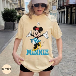 May include: Butter-colored t-shirt featuring a graphic of Minnie Mouse in a playful pose. Minnie is depicted in black, white, red, and yellow, with the name "MINNIE" in blue below. The shirt has short sleeves and a relaxed fit.