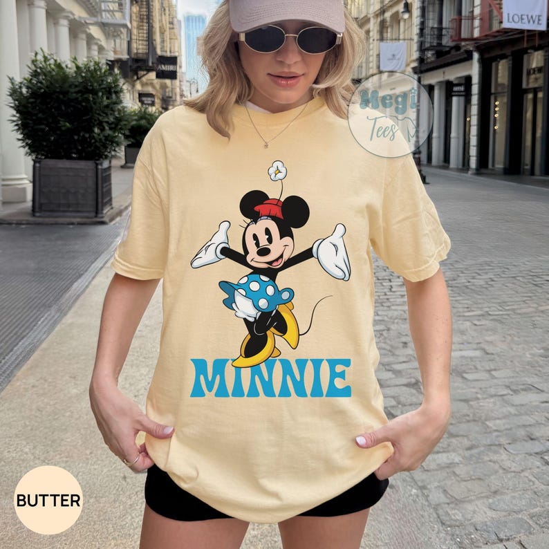 May include: Butter-colored t-shirt featuring a graphic of Minnie Mouse in a playful pose. Minnie is depicted in black, white, red, and yellow, with the name "MINNIE" in blue below. The shirt has short sleeves and a relaxed fit.