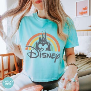 May include: A light blue t-shirt featuring a colorful rainbow design with a gray castle and the word "Disney" in a stylized font. The shirt has short sleeves and a crew neck. The text "Chalky Mint" is in the lower left corner.