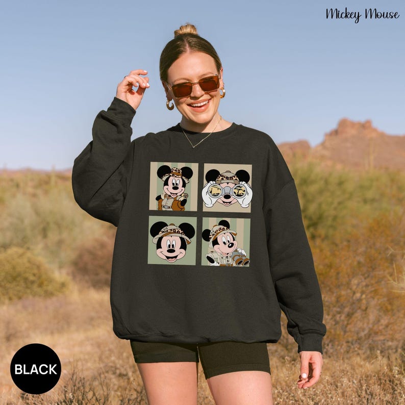 May include: Black sweatshirt featuring four panels with Mickey Mouse in safari attire. The panels are set against a green and beige striped background. The words "Mickey Mouse" are in the upper right corner, and the word "BLACK" is in a black circle at the bottom left.