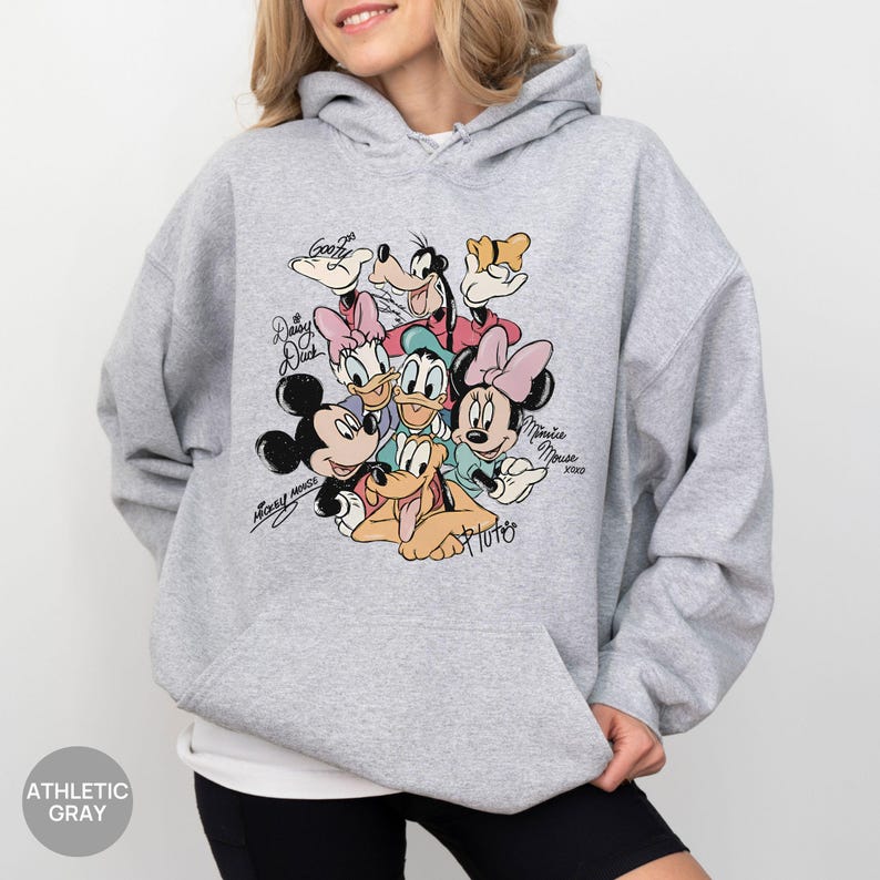 K&ouml;nnte beinhalten: Grauer Kapuzenpullover mit einem farbenfrohen Aufdruck von Disney-Figuren wie Mickey Mouse, Minnie Mouse, Donald Duck, Daisy Duck, Goofy und Pluto. Der Hoodie hat eine Kapuze mit Kordelzug und eine Vordertasche. Der Text "Athletic Gray" ist unten links aufgedruckt.
