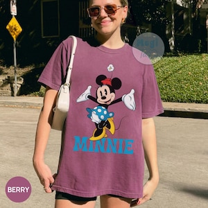 May include: Berry-colored t-shirt featuring a Minnie Mouse graphic. The design includes Minnie in a red hat and blue polka-dot skirt, with the word "MINNIE" in blue block letters. The shirt is a relaxed fit.