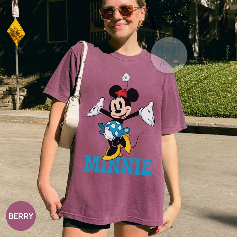 May include: Berry-colored t-shirt featuring a Minnie Mouse graphic. The design includes Minnie in a red hat and blue polka-dot skirt, with the word "MINNIE" in blue block letters. The shirt is a relaxed fit.