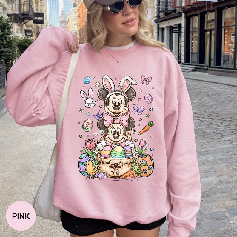 Puede incluir: Sudadera rosa claro con un dise&ntilde;o de Mickey y Minnie Mouse como conejos de Pascua, rodeados de huevos de Pascua, zanahorias y flores. La palabra "PINK" est&aacute; impresa en la parte inferior izquierda de la sudadera.