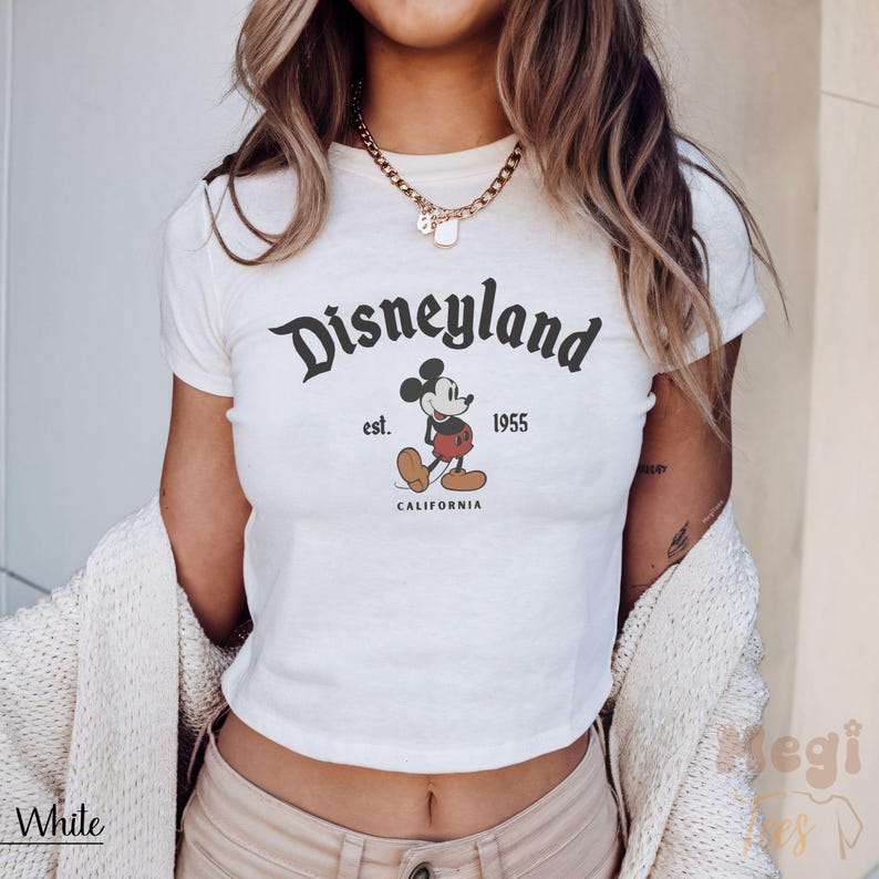 May include: White crop top featuring the Disneyland logo with Mickey Mouse. The design includes the text "Disneyland est. 1955 CALIFORNIA" in black. The shirt is a classic, casual style.