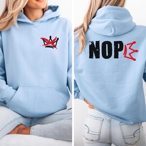 May include: Light blue hoodie with a front pocket and a hood. The front features a red and black crown design. The back of the hoodie displays the word "NOPE" in black with a red crown design. The hoodie is made of a soft material.