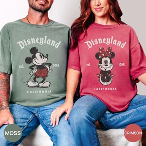 Puede incluir: Dos camisetas de manga corta, una verde musgo y otra carmes&iacute;, con el logotipo de Disneyland y Mickey y Minnie Mouse. Las camisetas tienen un dise&ntilde;o vintage con el texto "est. 1955 CALIFORNIA". Los modelos llevan vaqueros azules.